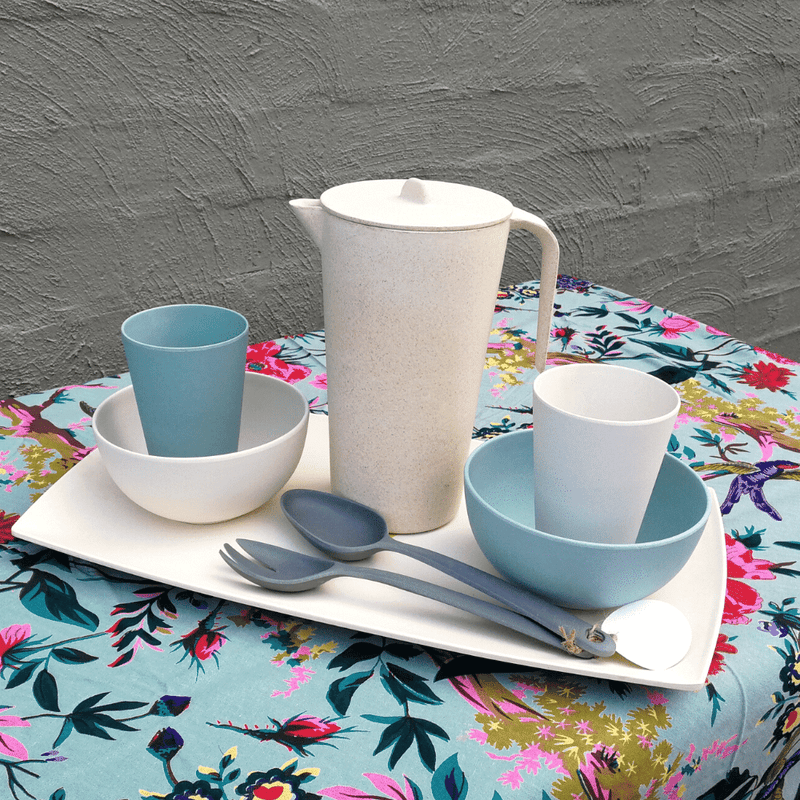 Bamboo Platter with Bamboo bowls in oat and sea colours, with bamboo cups inside in the same oat and sea colours and a bamboo jug sitting on the platter.  There are also some bamboo salad servers in charcoal, sitting on a blue tablecloth with pink flowers and birds 